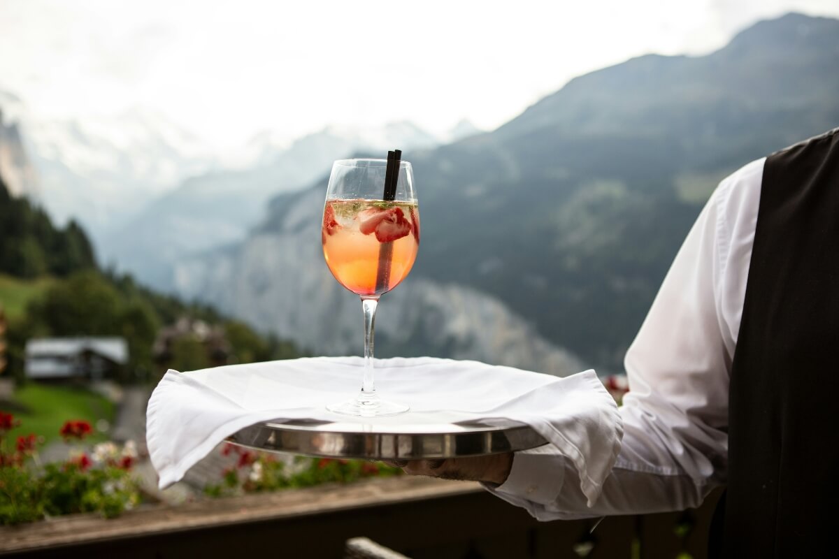 Orange spritz cocktail on server's tray with dramatic Alpine mountain landscape background