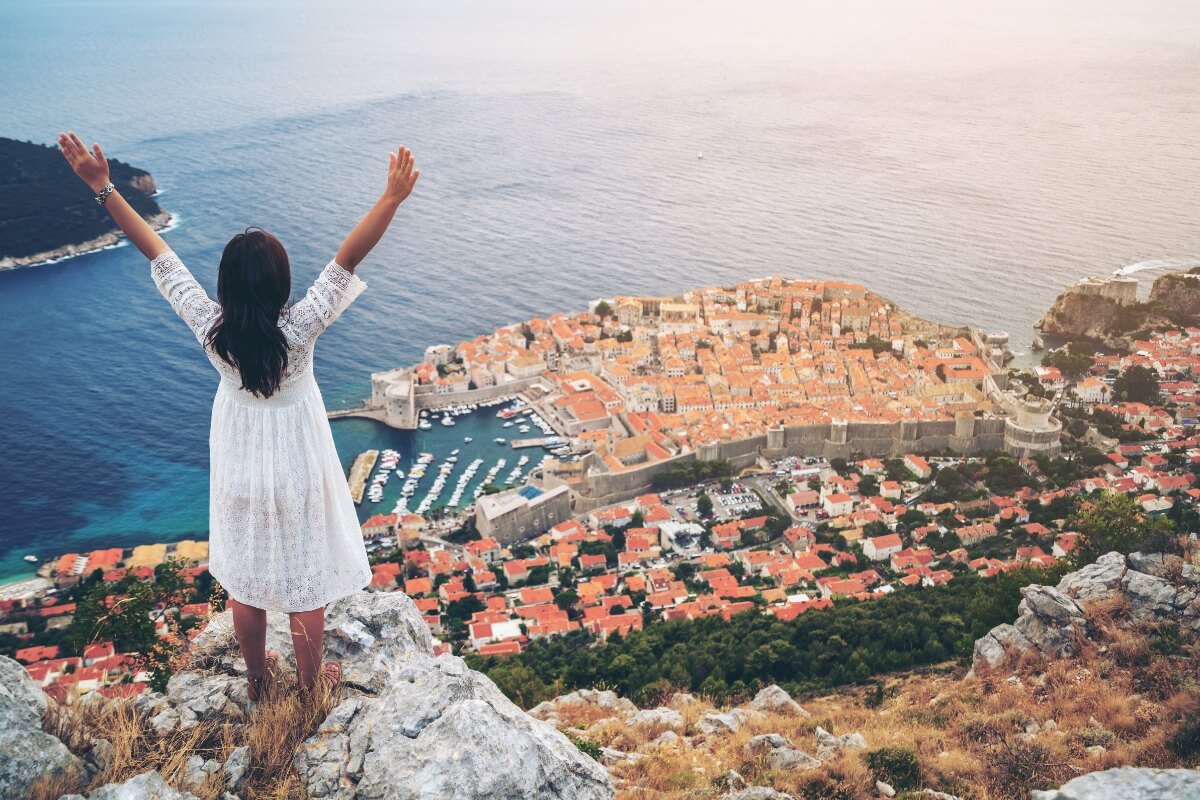 Woman in white dress with arms raised overlooking Dubrovnik's Old Town, Adriatic Sea, and terracotta rooftops