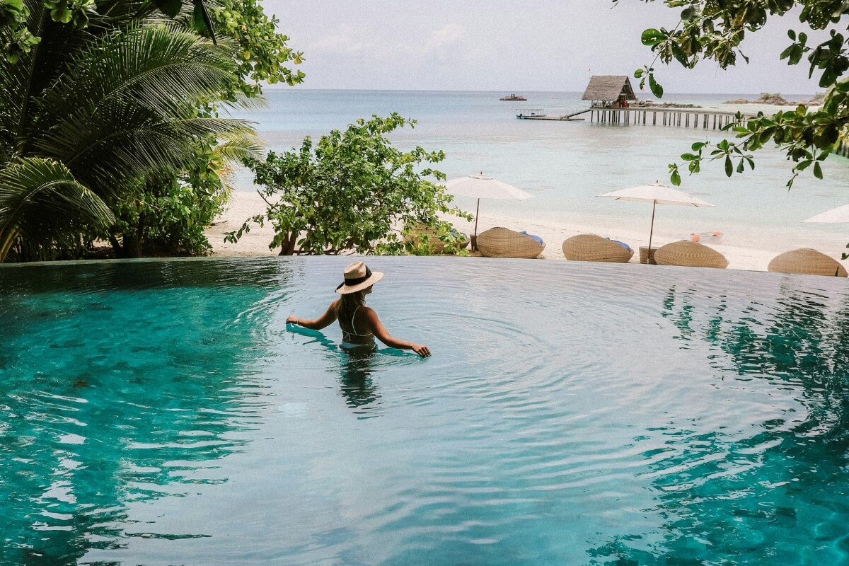 Person in infinity pool overlooking pristine white sand beach, turquoise ocean and overwater villa in tropical paradise