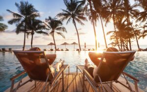 Two people relaxing on loungers by infinity pool overlooking ocean at sunset, silhouetted palm trees, tropical beach resort vacation