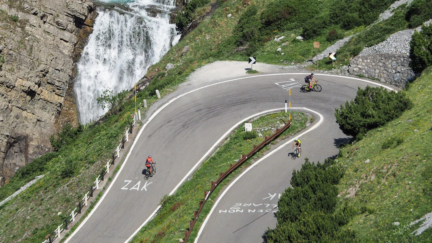 Cyclists navigating dramatic Alpine hairpin turns near cascading waterfall on Ride & Seek mountain tour