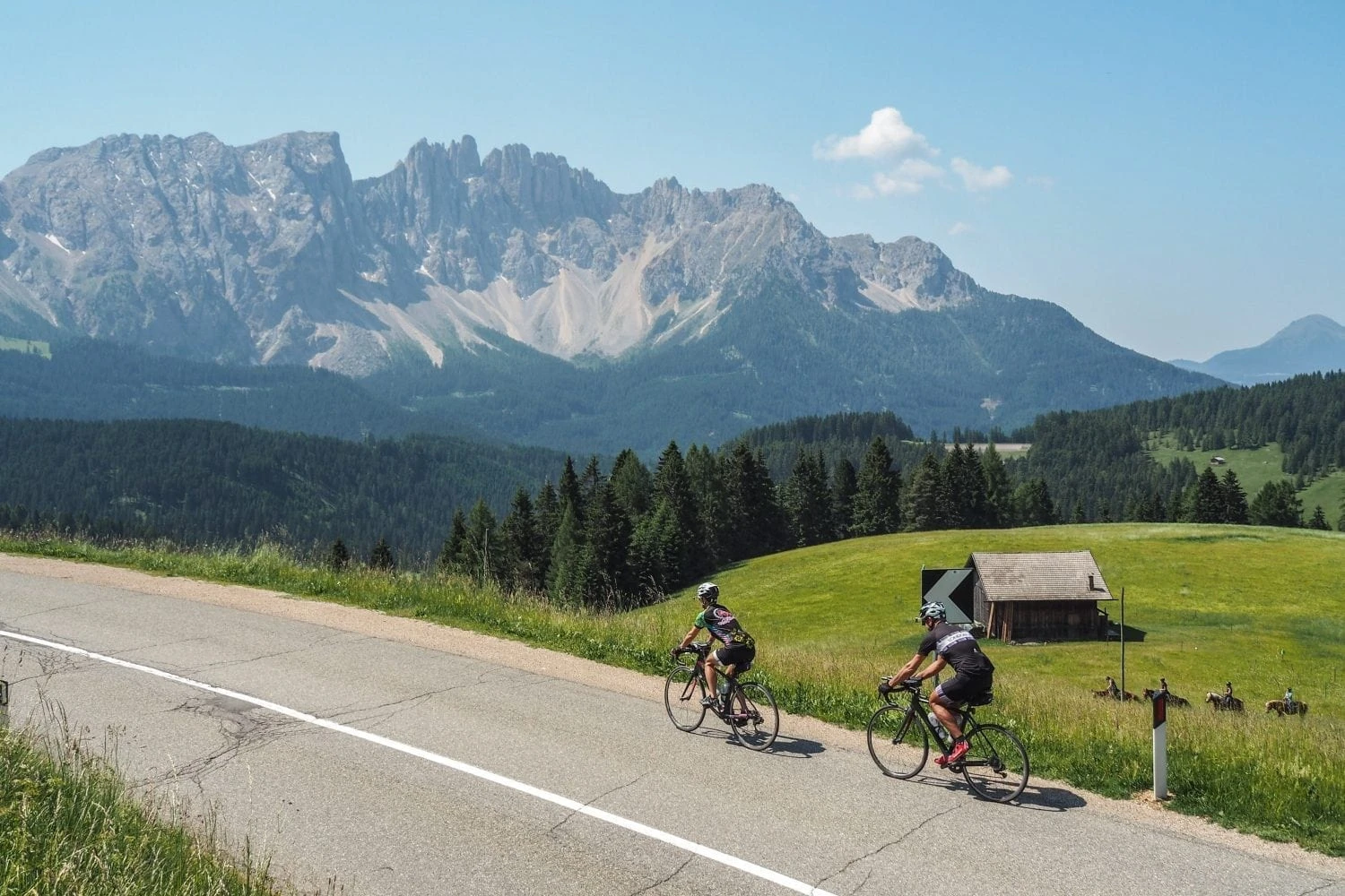 Two cyclists riding past wooden cabin with dramatic Dolomites mountain peaks on Ride & Seek tour