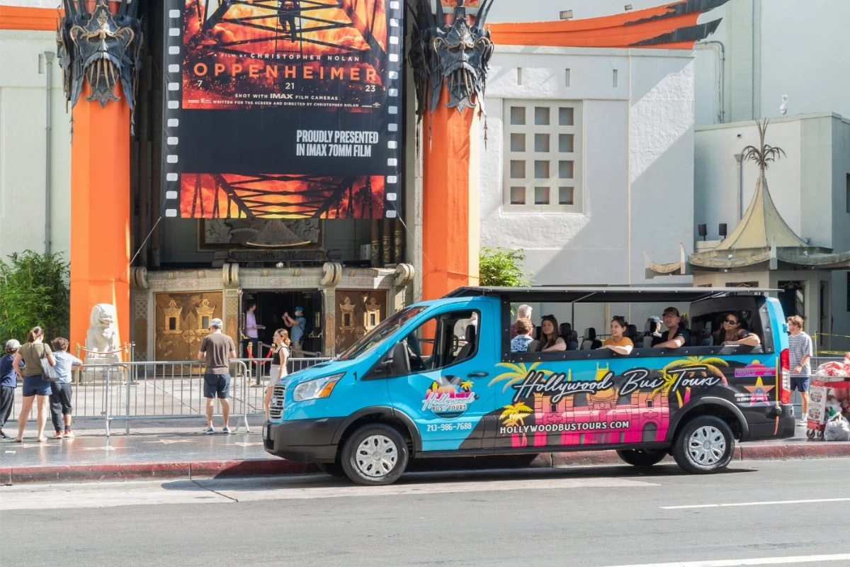 Colorful Hollywood bus tour vehicle parked outside iconic Chinese Theatre with Oppenheimer movie poster and tourists nearby
