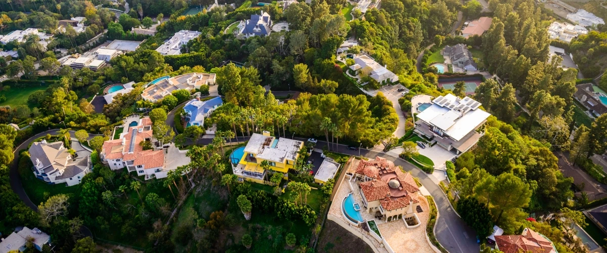 Aerial view of luxury mansions with pools in the Hollywood Hills neighborhood of Los Angeles, showcasing Spanish and Mediterranean architecture surrounded by lush greenery and winding streets during a celebrity homes tour.