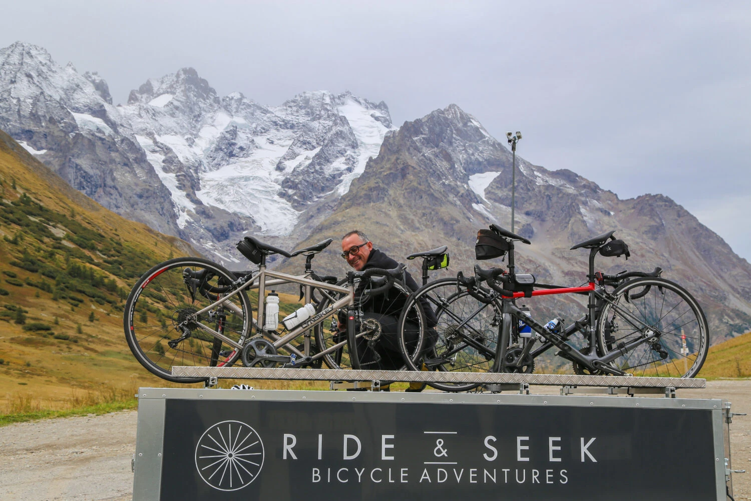 Ride & Seek branded support vehicle with bikes mounted against dramatic snow-capped mountain backdrop