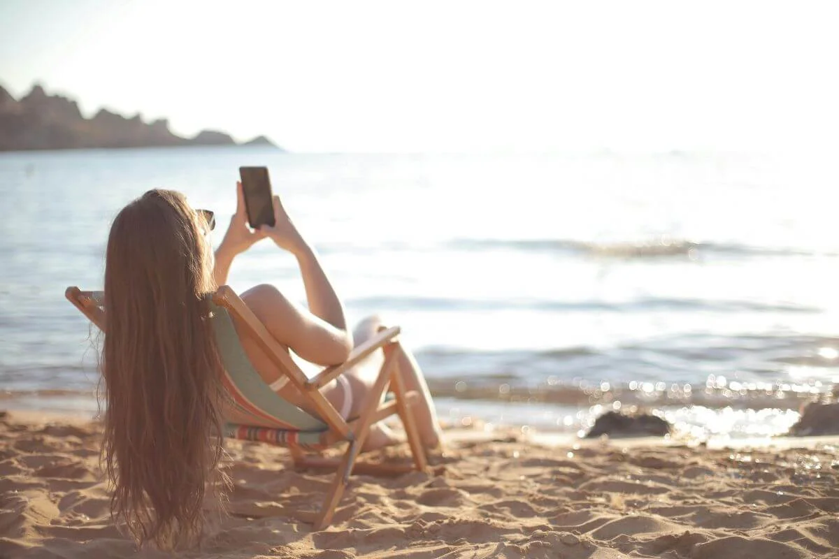 A traveler relaxing on a beach chair while using a smartphone to capture and share a seaside view, symbolizing how travel brands can increase AI visibility by showing up where travelers search, discover, and share experiences online.