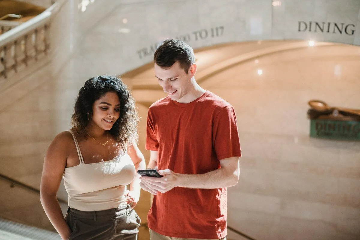 Two people standing indoors looking at a smartphone together, suggesting travelers using mobile search to plan dining or activities.
