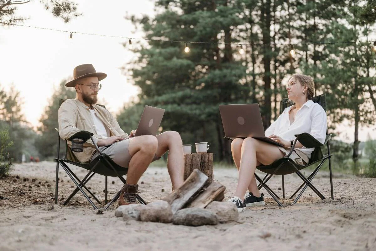 Two people sitting outdoors by a campfire, working on laptops in folding chairs, representing flexible remote work and location-independent collaboration.