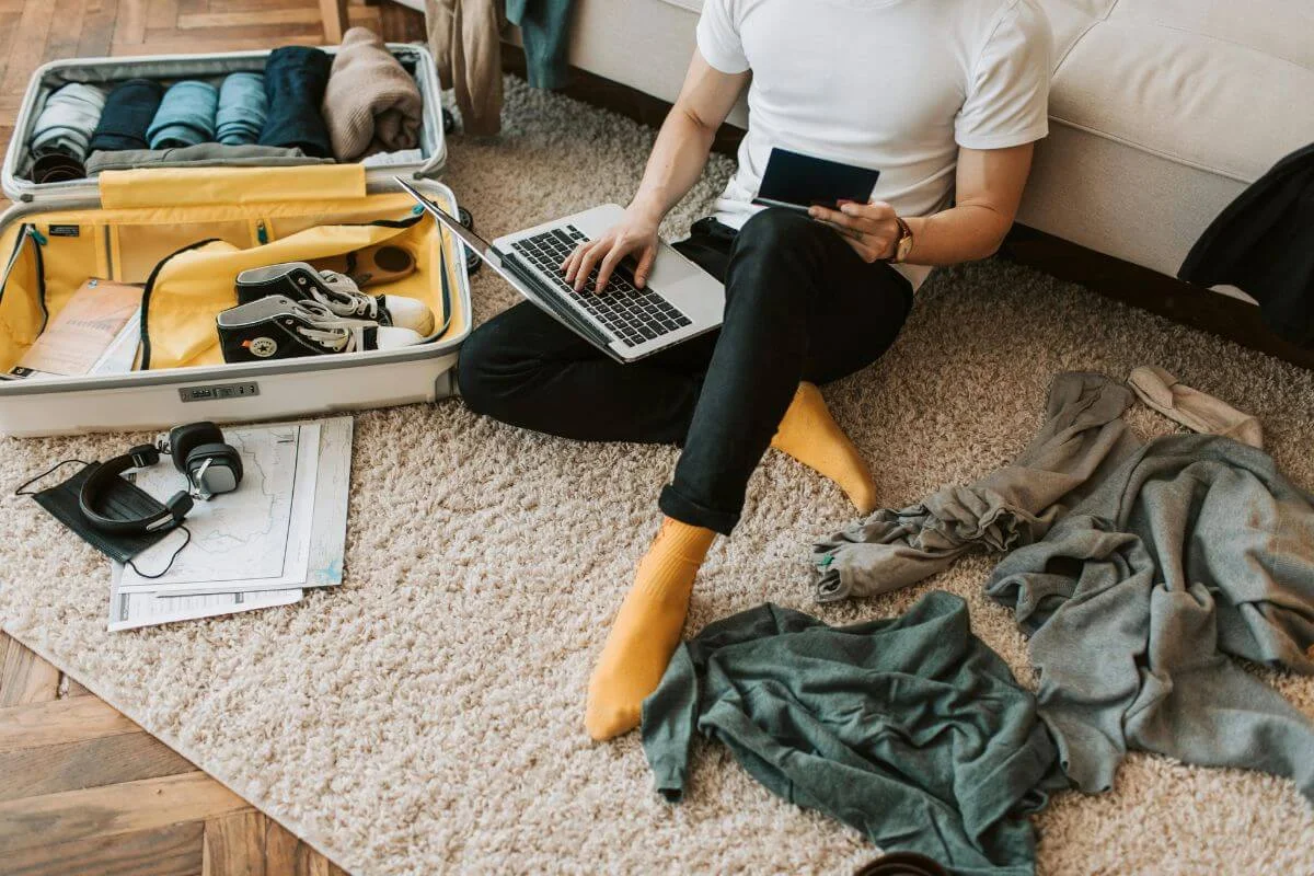 Person sitting on the floor with an open suitcase, using a laptop and holding a passport, showing travel planning and booking while packing for a trip.