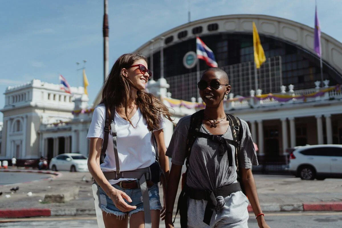  Two travelers wearing backpacks walk and smile in front of a large historic building with flags, exploring a city street during the day.