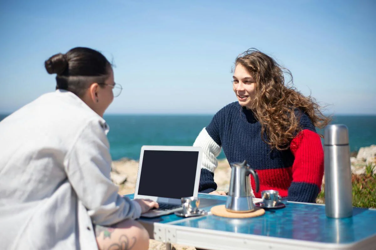  Two people sit at an outdoor table near the ocean, talking while using a laptop and enjoying coffee with a sea view in the background.