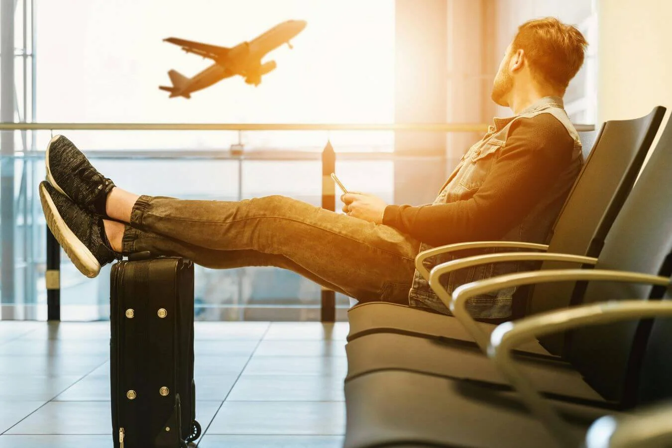Traveler sitting in an airport terminal with luggage, looking at a smartphone as an airplane takes off in the background