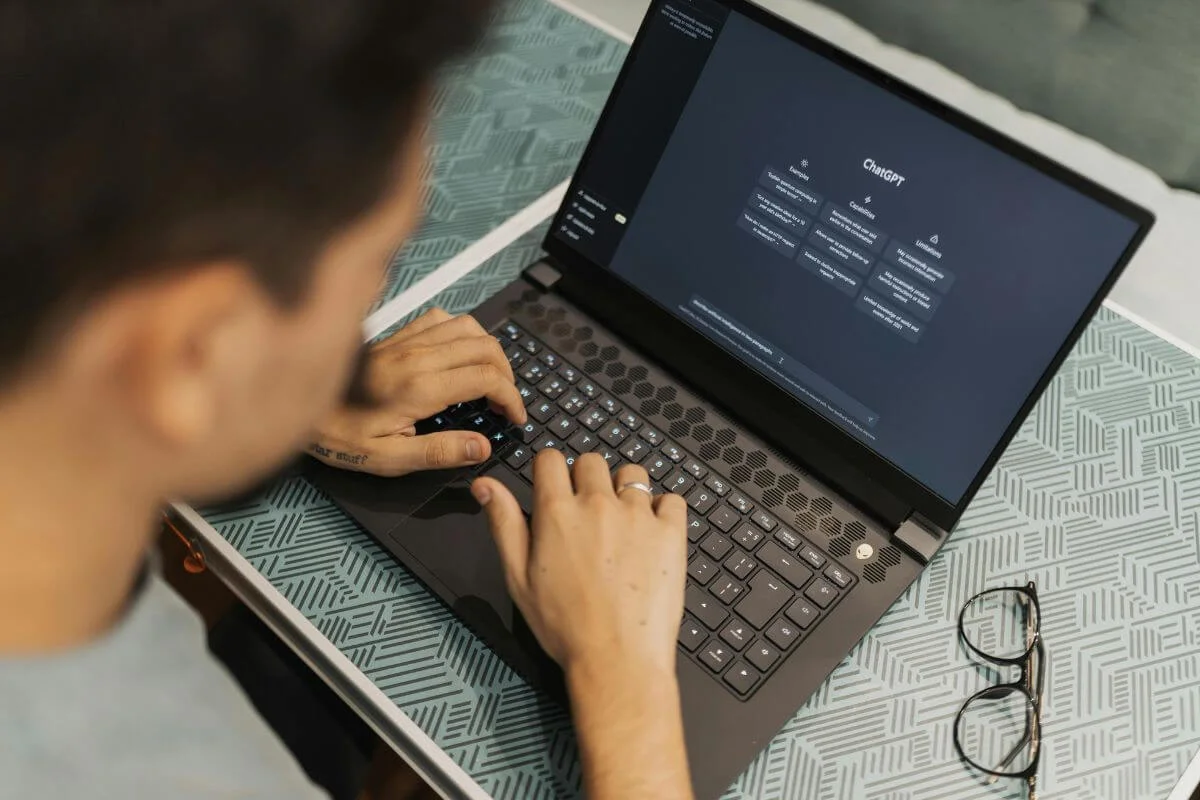  Person typing on a laptop displaying the ChatGPT interface on the screen, with a pair of glasses resting on the table beside the computer.