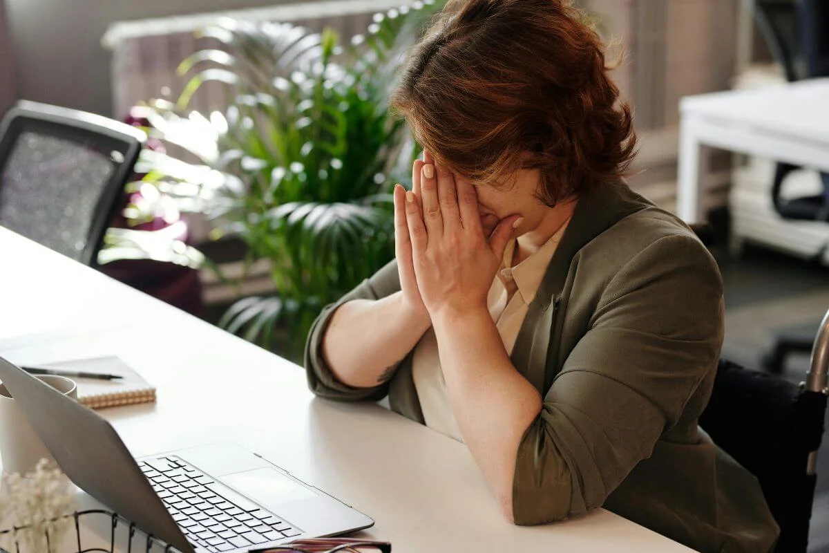  Person sitting at a desk with a laptop, covering their face with their hands, appearing stressed or overwhelmed in a home or office workspace.