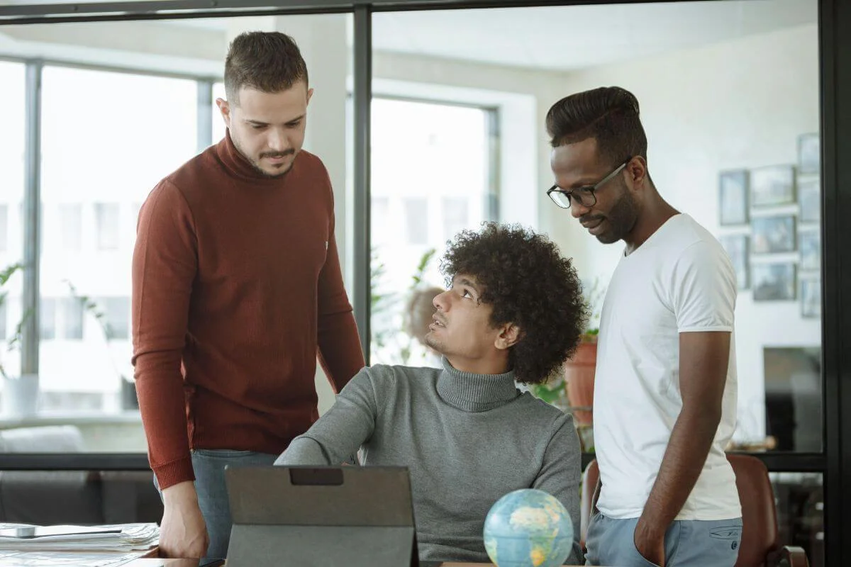  Three coworkers standing and sitting together in an office, discussing something on a laptop during a collaborative work session.