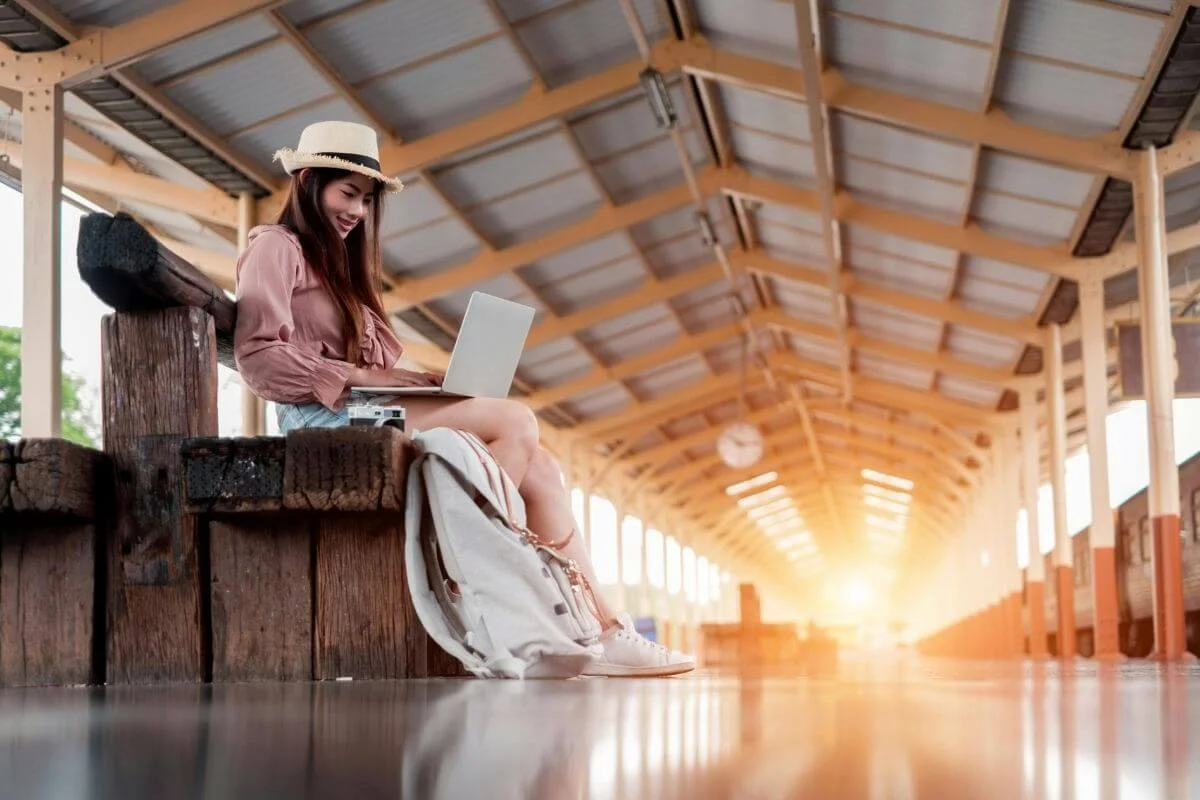 Woman sitting at a train station using a laptop, with a backpack beside her, working or planning travel while waiting for transportation.